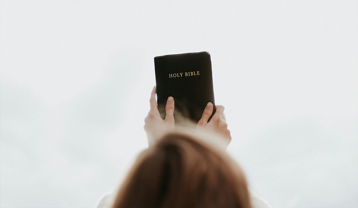 A woman holds up a black Bible against an overcast sky.