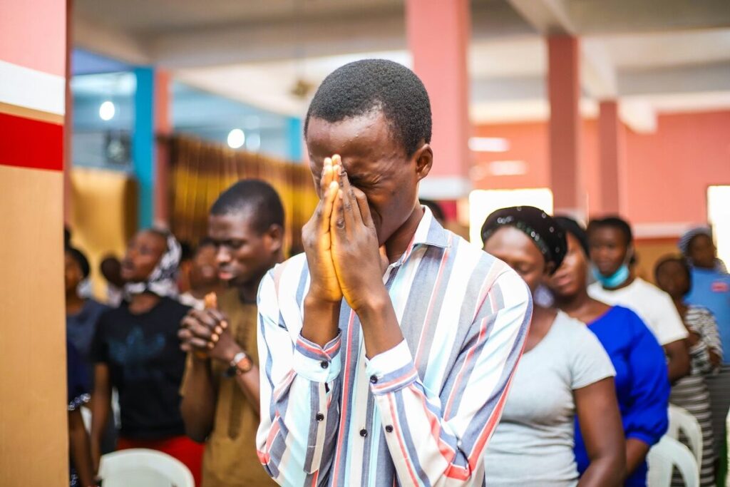 A man stands with his hands pressed together in front of his face, eyes closed, surrounded by others in a room, appearing to be in prayer or deep reflection.