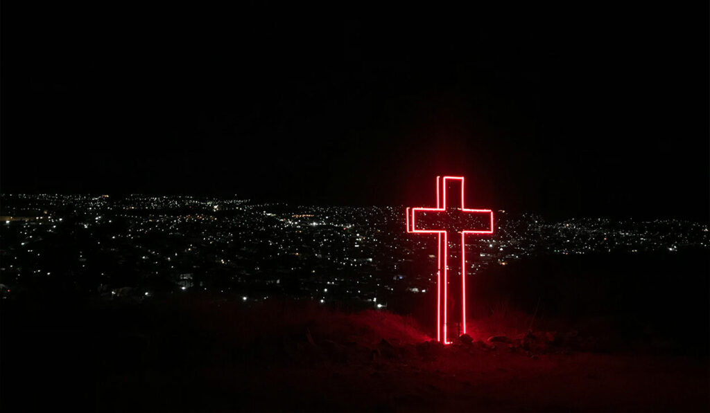 A cross illuminated with red, neon lights stands on top of a hill, overlooking a city at night.
