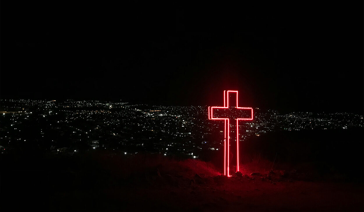 A cross illuminated with red, neon lights stands on top of a hill, overlooking a city at night.