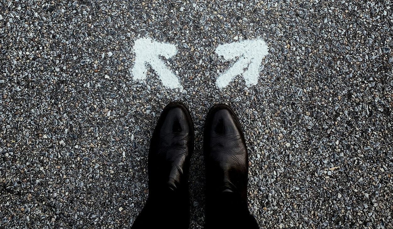 Two white arrows are painted on an asphalt pavement. A person wearing black dress shoes and slacks stands behind both arrows.