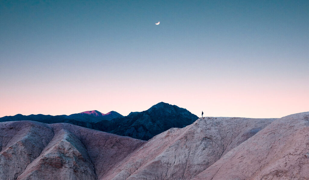 A person stands on a mountain range. A crescent moon is in the sky.