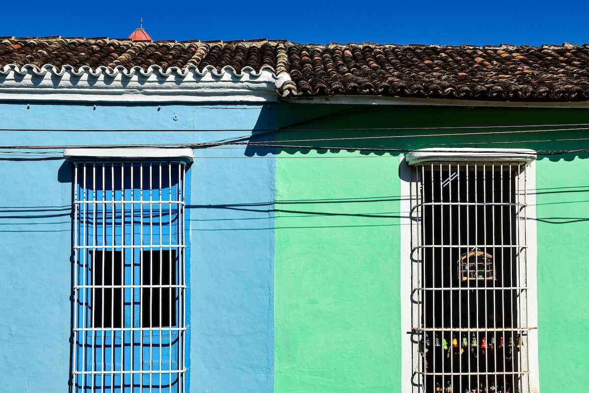 A building with blue and green walls, each featuring a barred window and intersected by several overhead wires, under a clear blue sky.