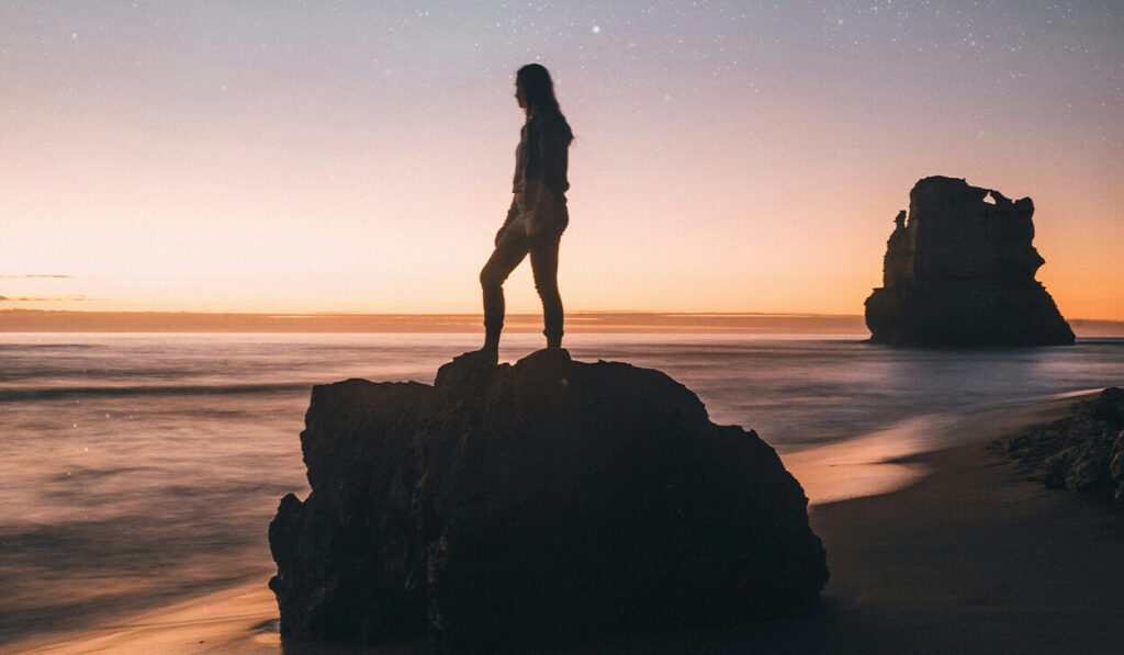 A woman stands on a boulder on a beach and looks out at the ocean. It's sunset, and there are stars in the sky.