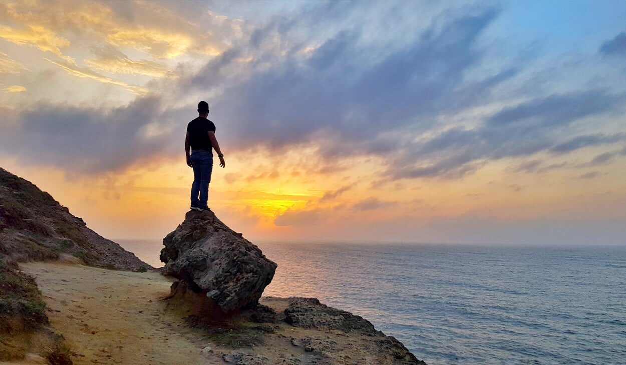 A man stands on a boulder on a beach and looks out at the ocean during sunrise.