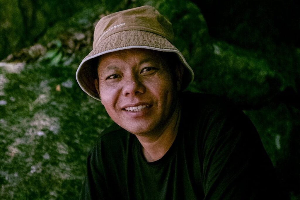 A person wearing a tan bucket hat and a dark shirt smiles outdoors with a natural, rocky background.