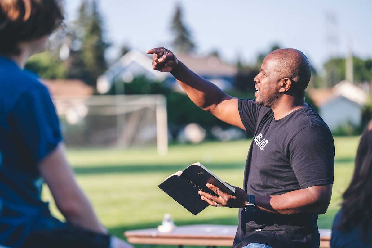A man holding an open book gestures while speaking outdoors, with blurred people and a soccer goal in the background.