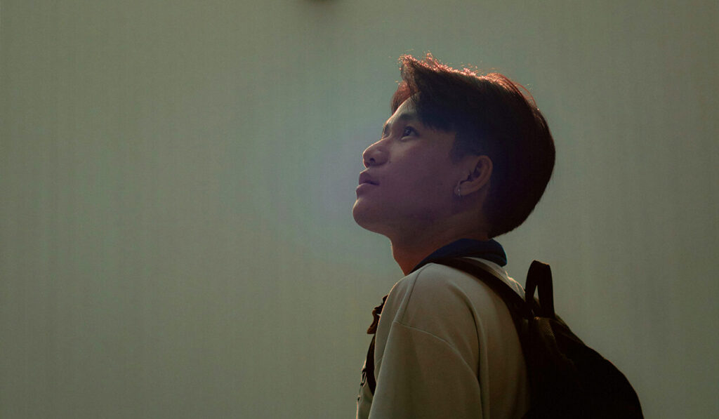 A man with a backpack looks at the ceiling in a plain room.