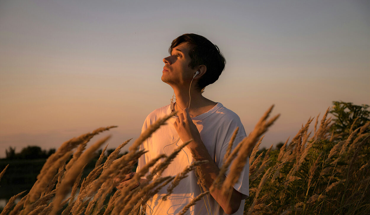 A man in a wheat field looks up at the sky. He is wearing a white t-shirt and has earbuds in his ears.