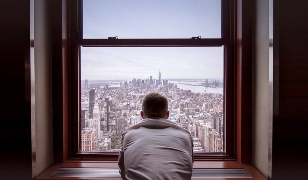 A man in a white t-shirt looks out a window over a skyline.