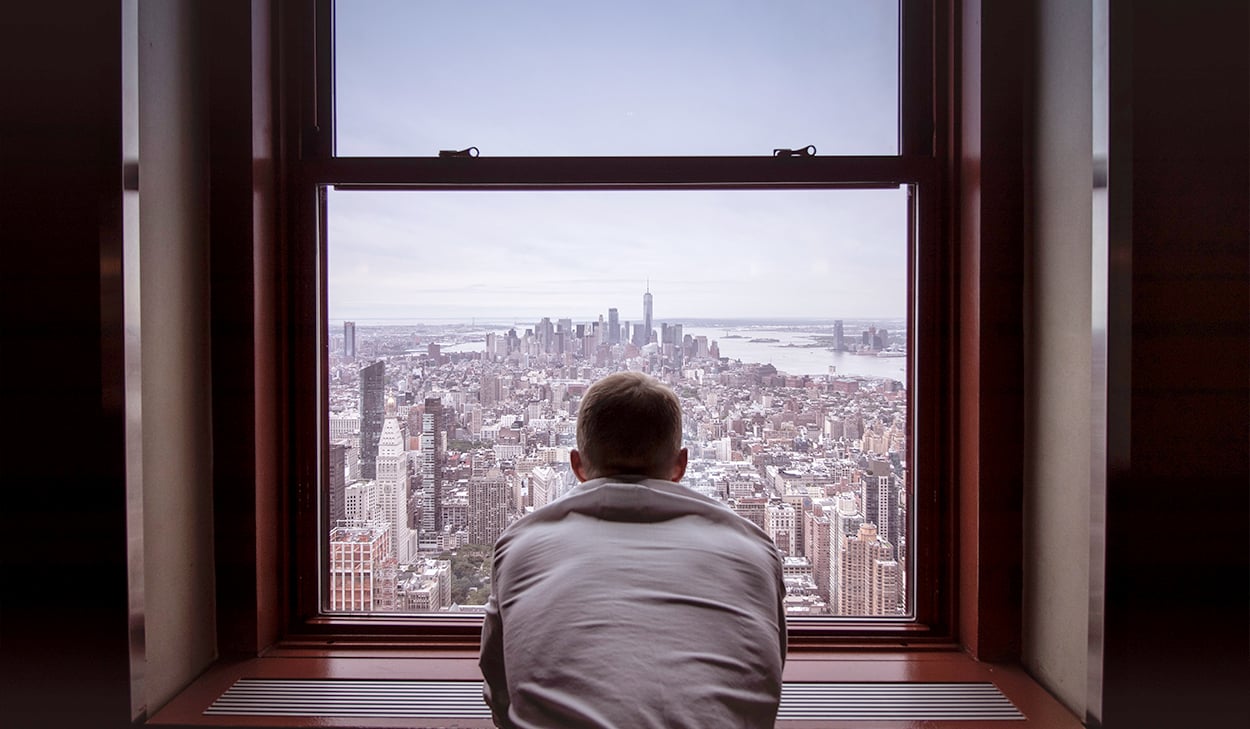 A man in a white t-shirt looks out a window over a skyline.
