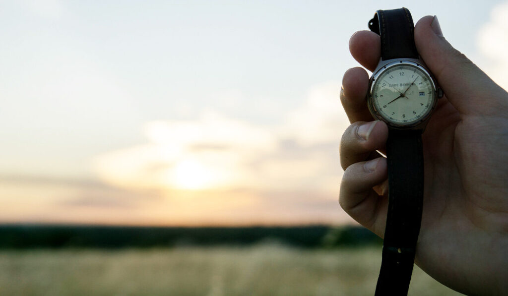 A person holds up a watch. A rising run is in the background.