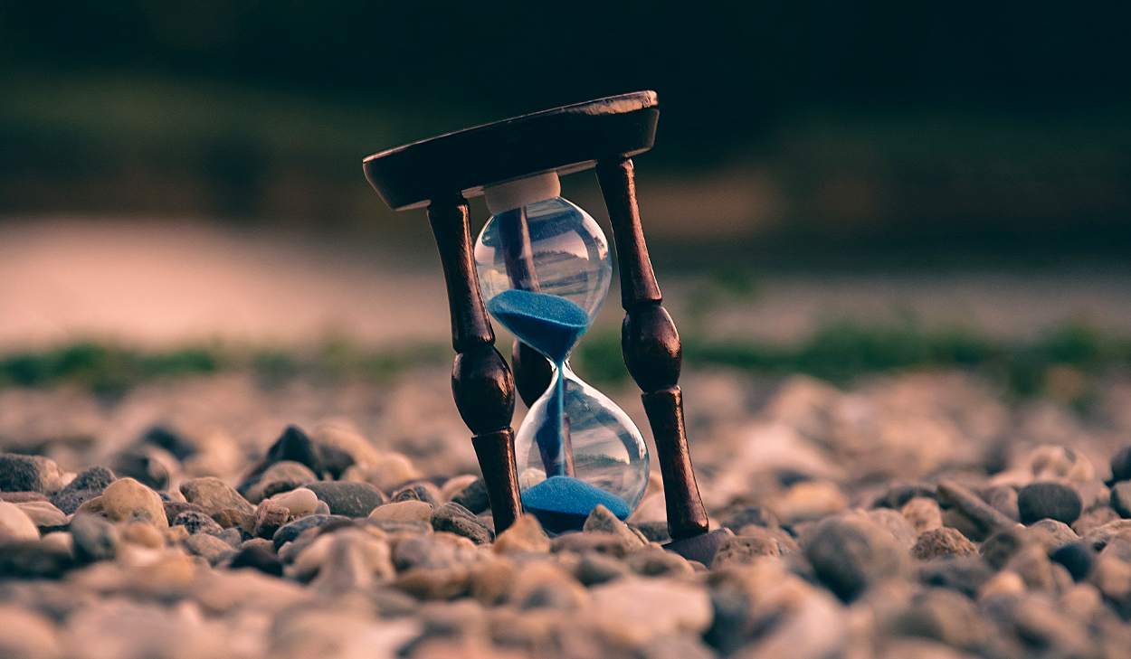 An hourglass with blue sand sits on rocky ground.