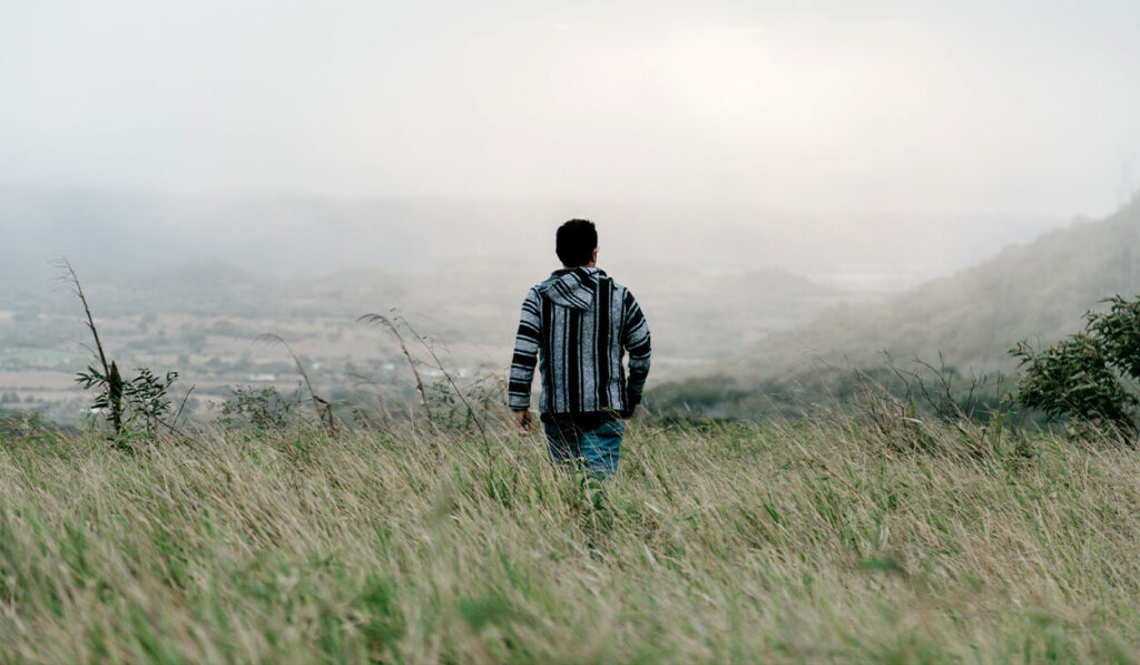 A man in a striped sweater walked through a grassy field. Fog is seen in the distance.