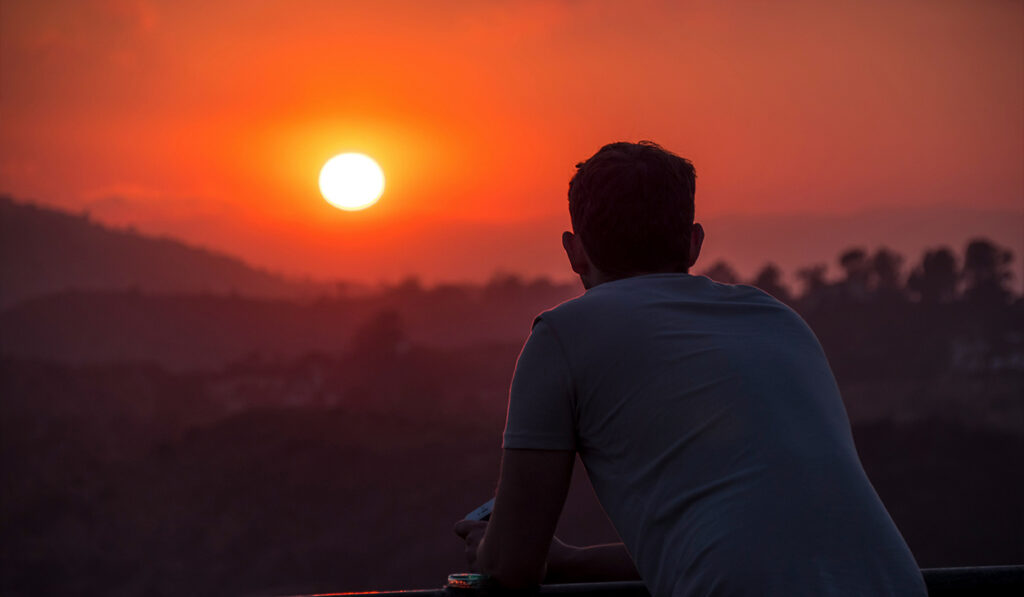 A man in a white shirt leans over a railing to look at the sunset.