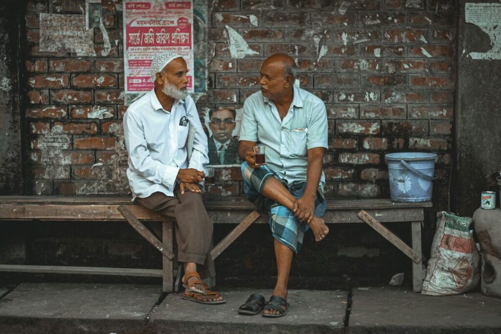 Two elderly men sit on a wooden bench against a weathered brick wall, engaged in conversation, with posters and various items in the background.