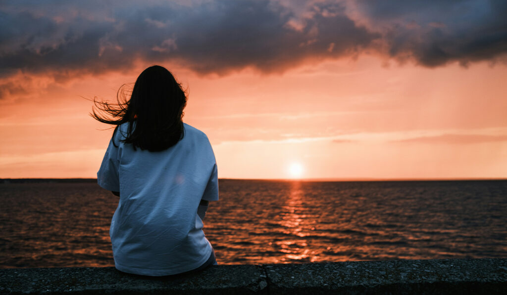 A woman in a white shirt looks at the sunset over a body of water.