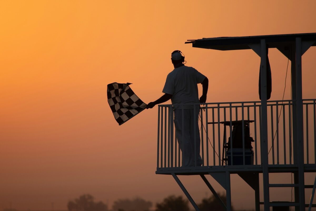 A person waves a checkered flag from a raised platform at sunset, silhouetted against an orange sky.