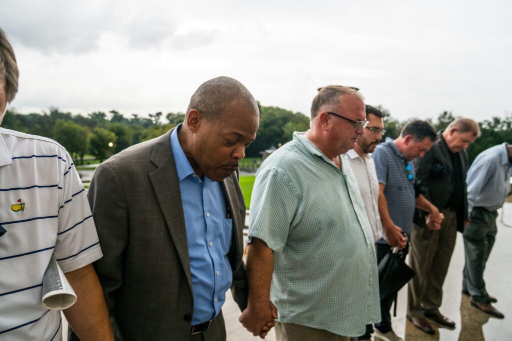 A group of people stand outdoors in a line, holding hands and bowing their heads in a moment of reflection or prayer.