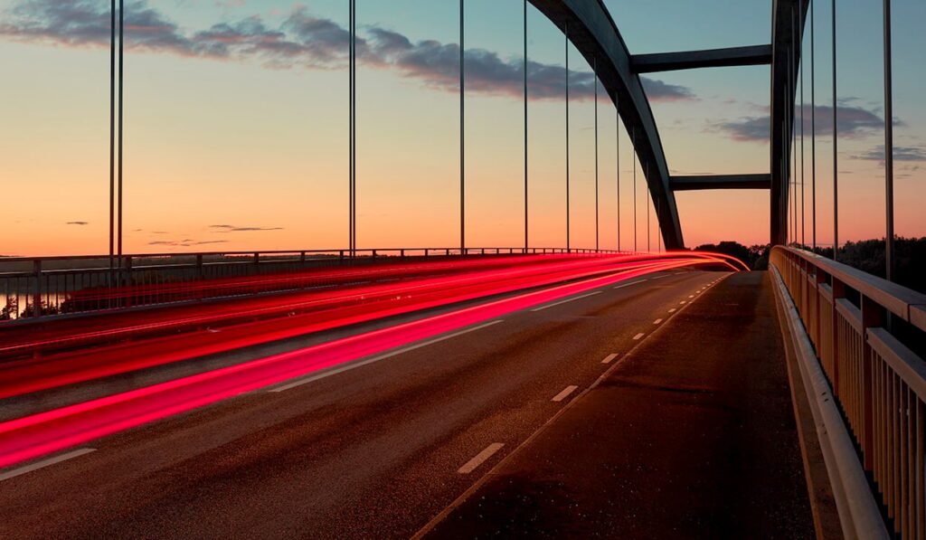 A long exposure image of tail lights on a bridge at sunset.