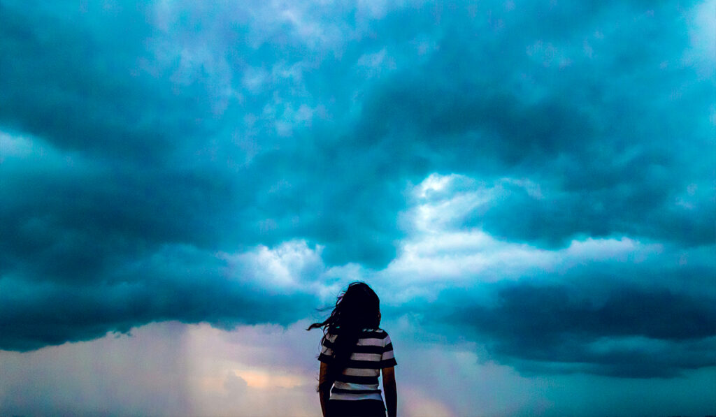 A woman in a black and white striped shirt stands facing away from the camera. Her hair is blowing in the wind while she looks up at the clouds.
