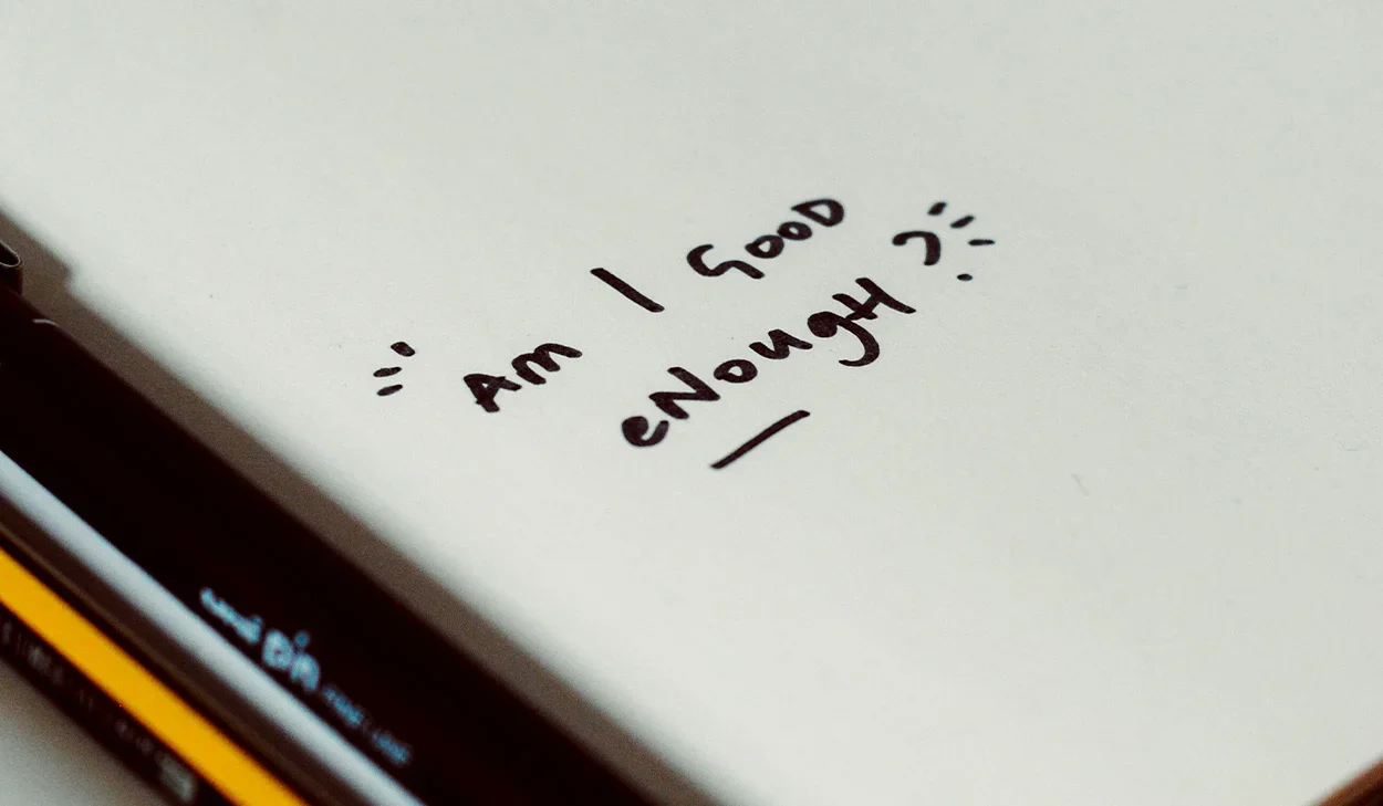 A sheet of white paper with the handwritten text Am I good enough? next to a black and yellow pen.