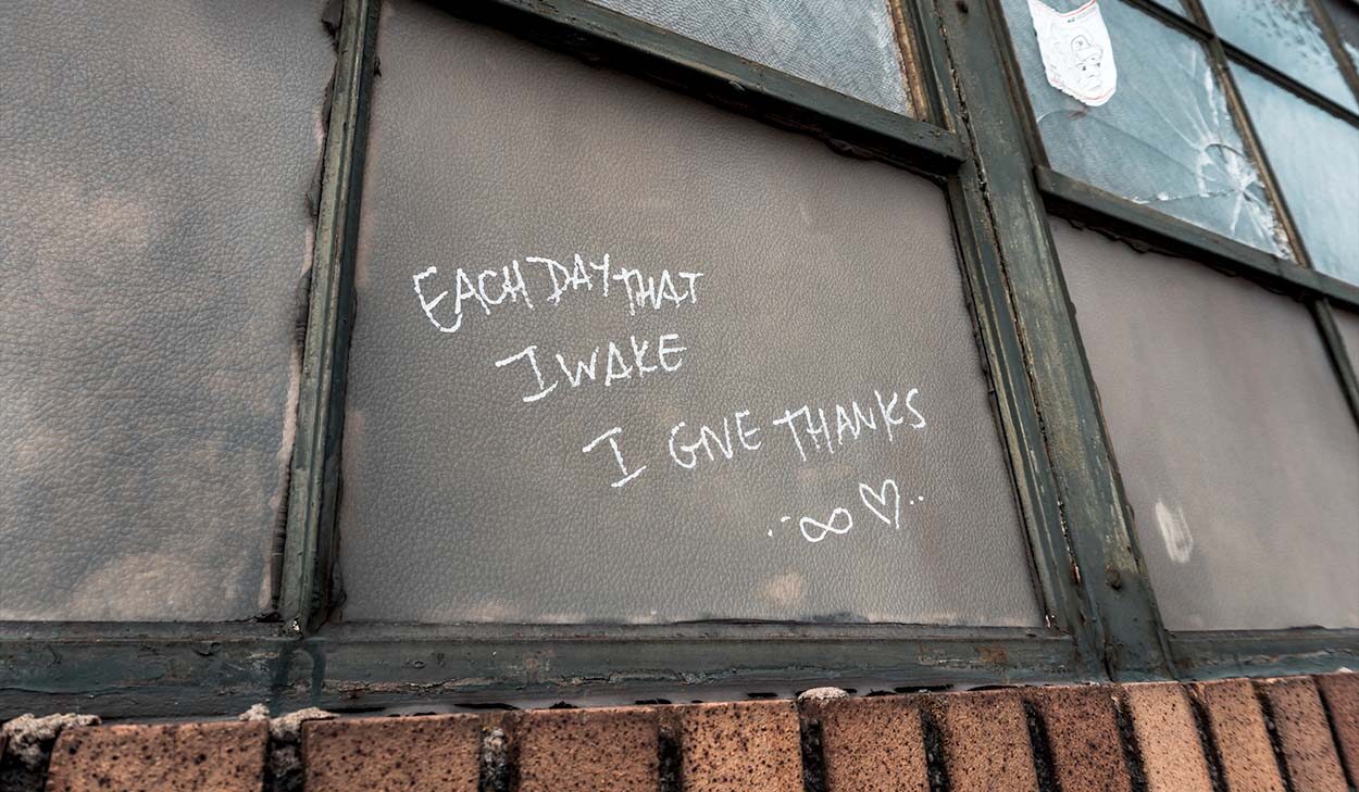 Graffiti on a weathered window reads Each day that I wake I give thanks ∞ ♥ in white chalk above a brick ledge.