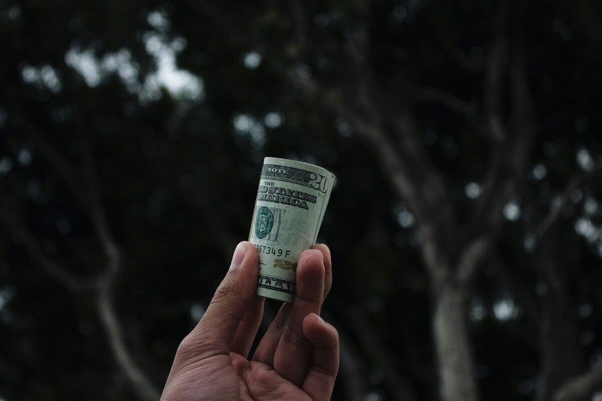 A hand holds a rolled-up U.S. dollar bill outdoors, with blurred trees in the background.