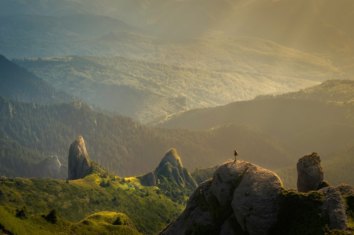 A person stands on a rocky peak overlooking a vast, sunlit valley with rolling hills and scattered rock formations.