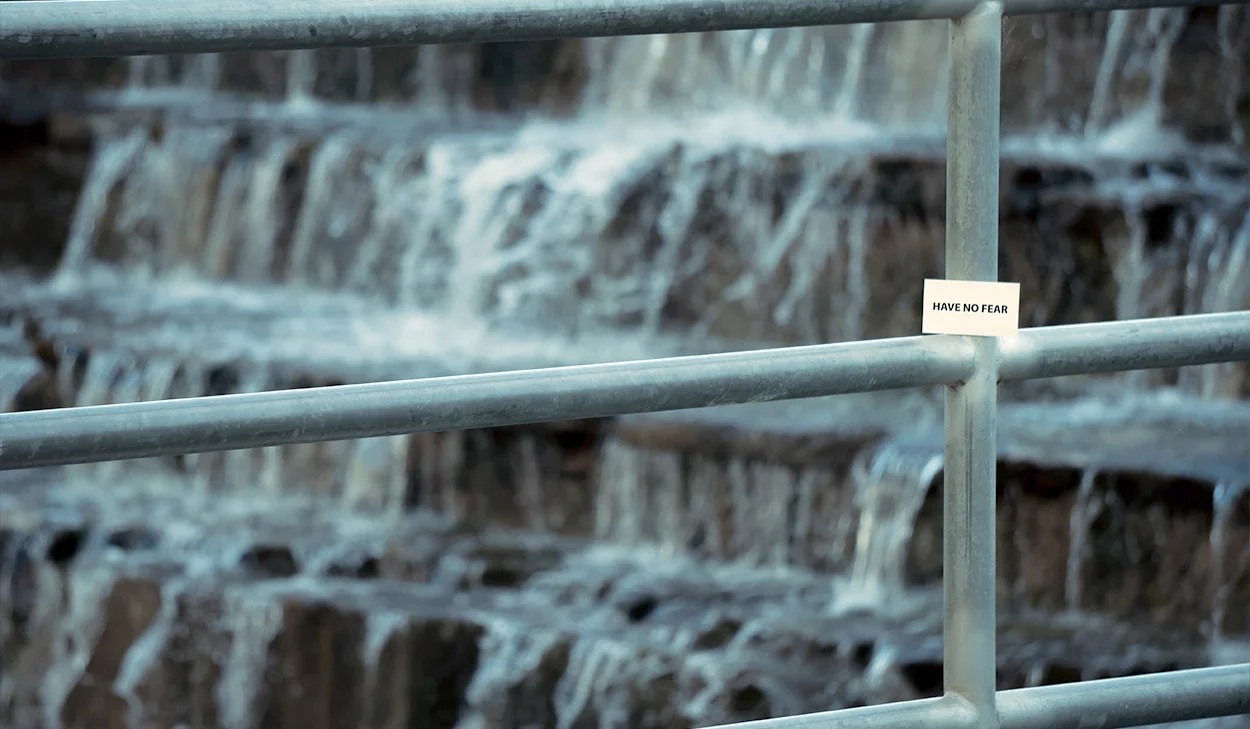 Metal railings in front of a waterfall with a small white sticker on the railing that reads HAVE NO FEAR.