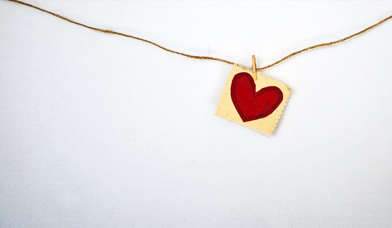 A square piece of paper with a red heart drawn on it hangs from a string using a clothespin against a plain white background.