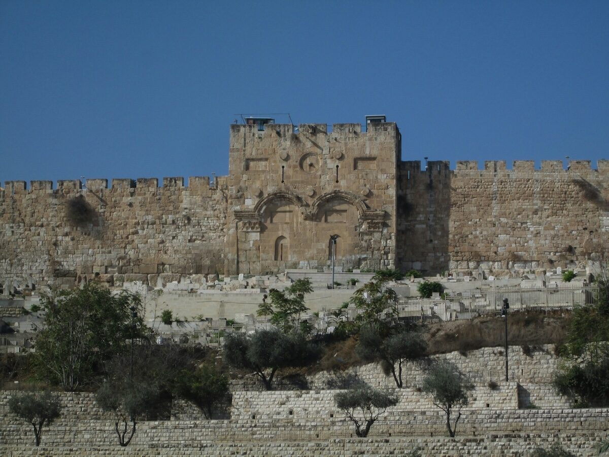 Stone walls and the sealed Golden Gate of Jerusalem’s Old City are shown under a clear blue sky, with trees and terraced stonework in the foreground.