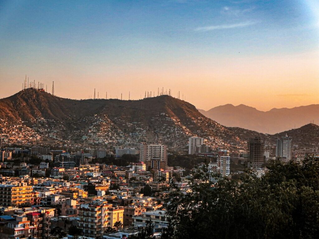 Cityscape at sunset with densely packed buildings in the foreground and hills with numerous antennas in the background under a clear sky.