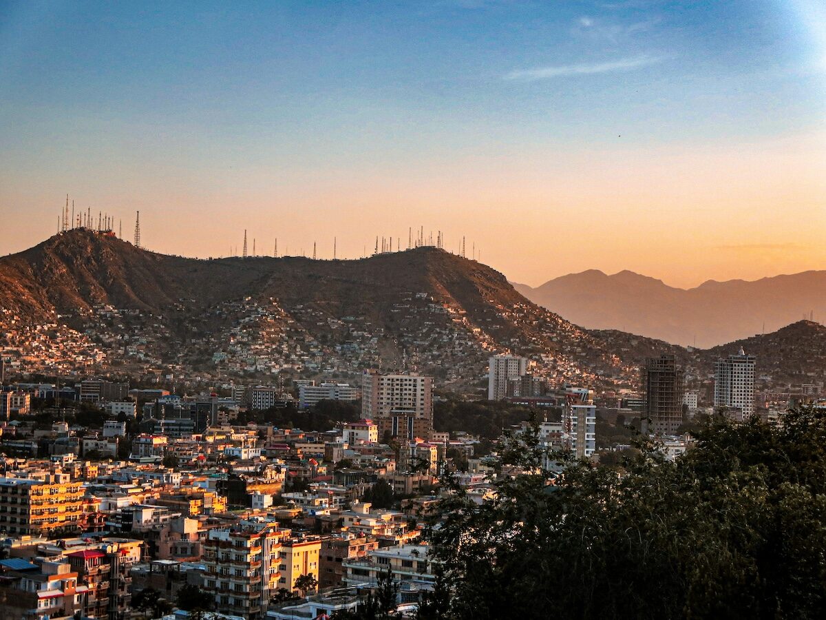 Cityscape at sunset with densely packed buildings in the foreground and hills with numerous antennas in the background under a clear sky.