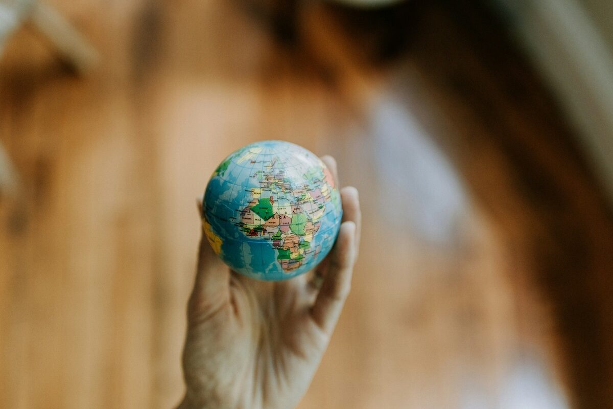 A hand holds a small globe, showing parts of Europe, Africa, and Asia, with a blurred wooden floor in the background.