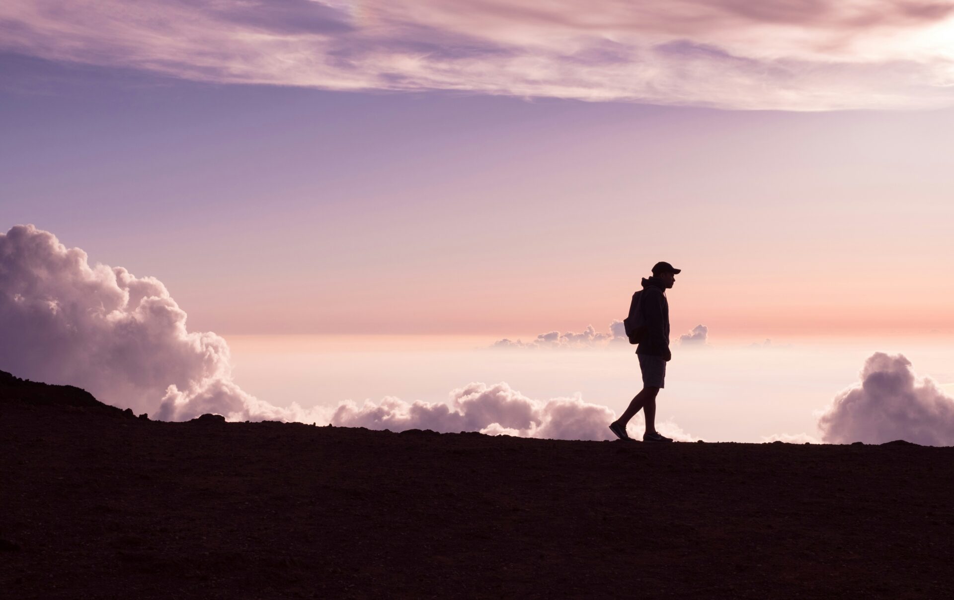 A person with a backpack walks along a ridge at sunset, with clouds and a colorful sky in the background.