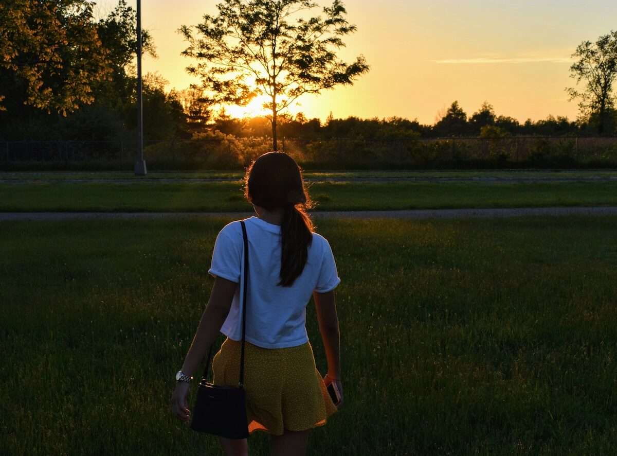 A person with a ponytail, wearing a white shirt and yellow shorts, walks through a grassy field at sunset.