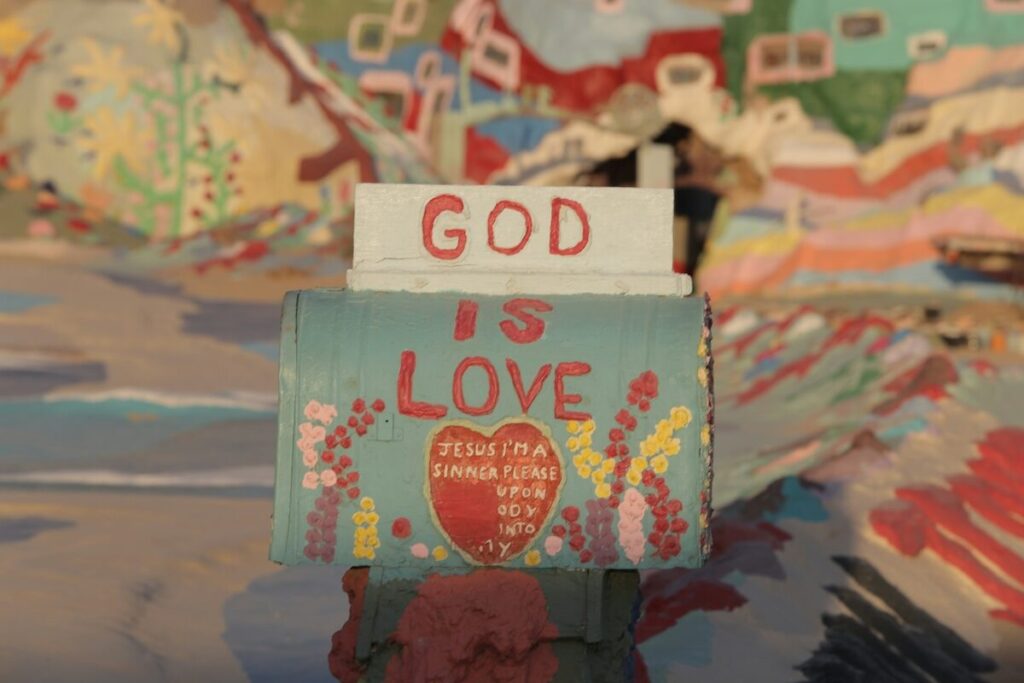 A mailbox painted with the words God is Love and colorful designs, set against a brightly painted, abstract background.