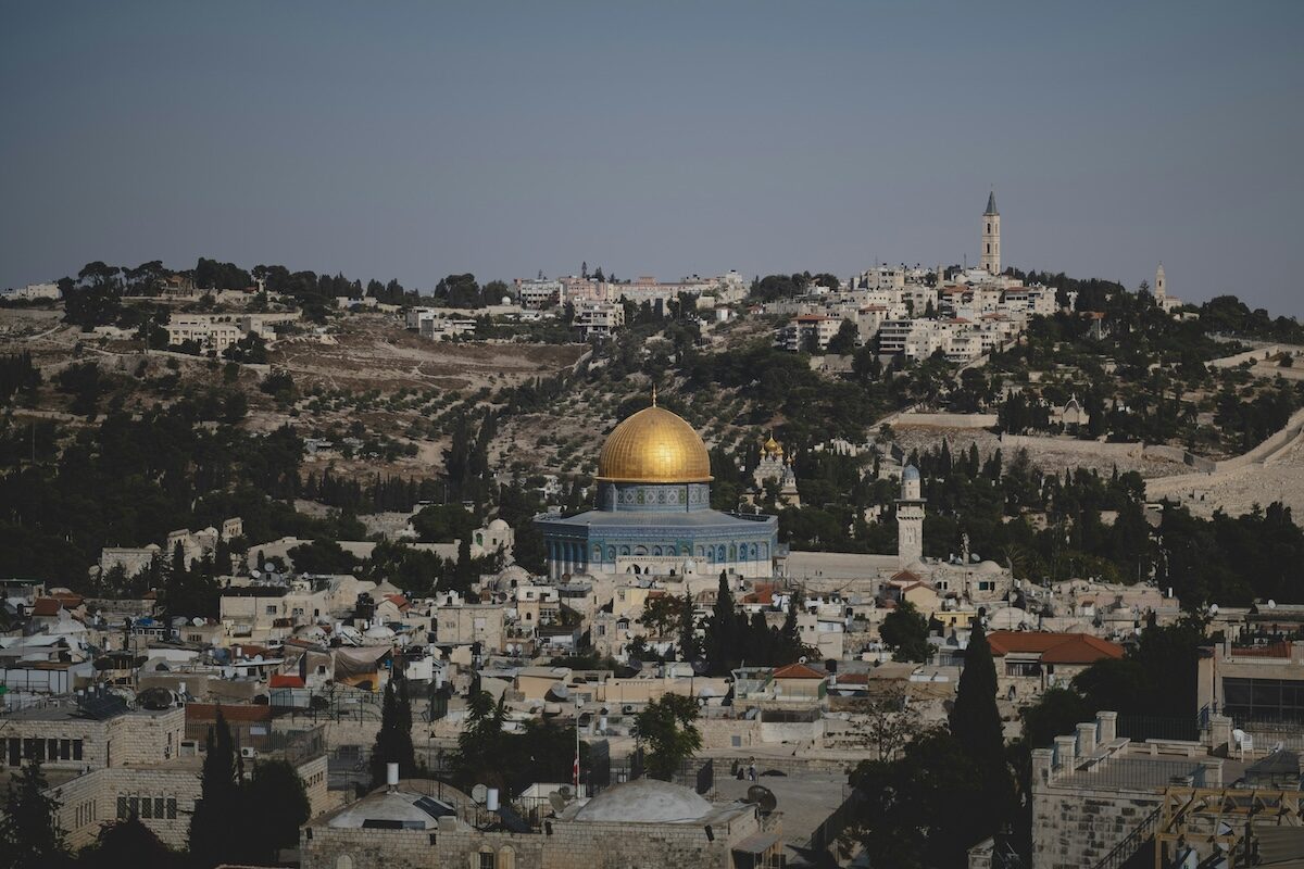 A view of Jerusalem showing the Dome of the Rock with its golden dome surrounded by buildings and hills in the background under a clear sky.