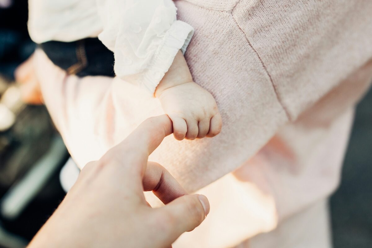 An adult hand reaches out and touches the hand of a baby who is wearing a light pink outfit.