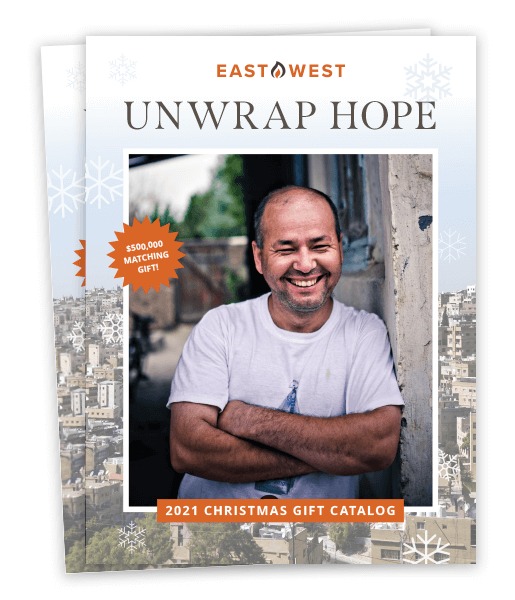 East-West branded catalog showing a bald, light-skinned man smiling and crossing his arms while leaning against a building. Above the photo, orange and black text reads 'East West; Unwrap Hope'. An orange badge over part of the photo reads '$500,000 matching gift!'. White text on an orange background underneath the photo reads '2021 Christmas gift catalog'.