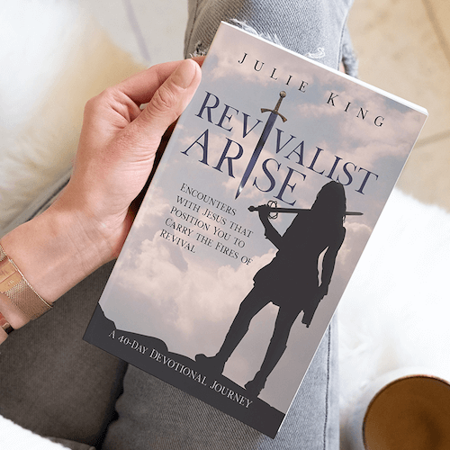 Close up of a white person's hand holding a book titled 'Revitalist Arise'. The book cover shows a cloudy sky with a silhouette of a woman holding a sword over one shoulder.