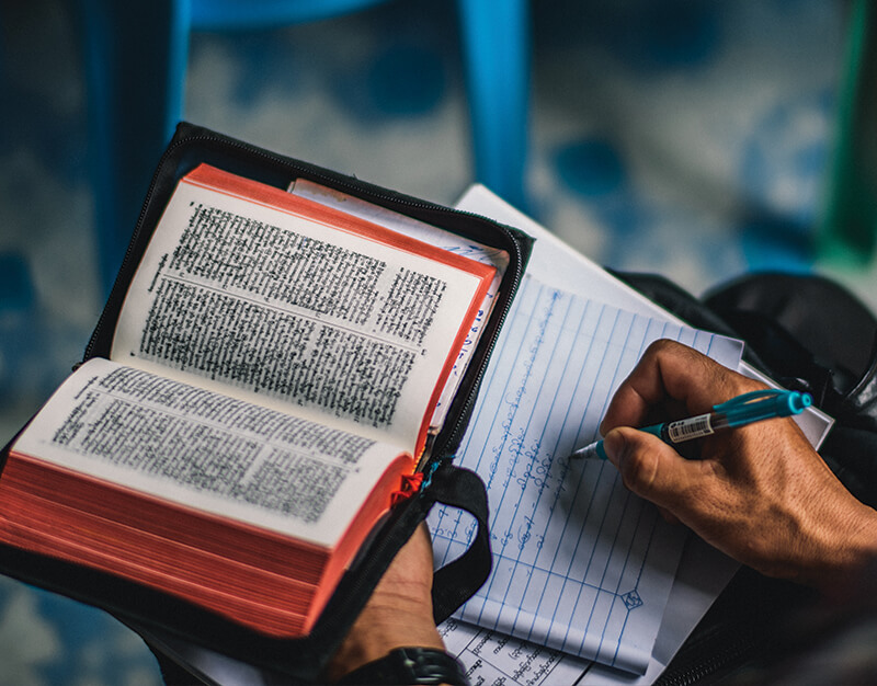 Close up of a person holding a Bible in one hand and writing in a notebook with the other hand.