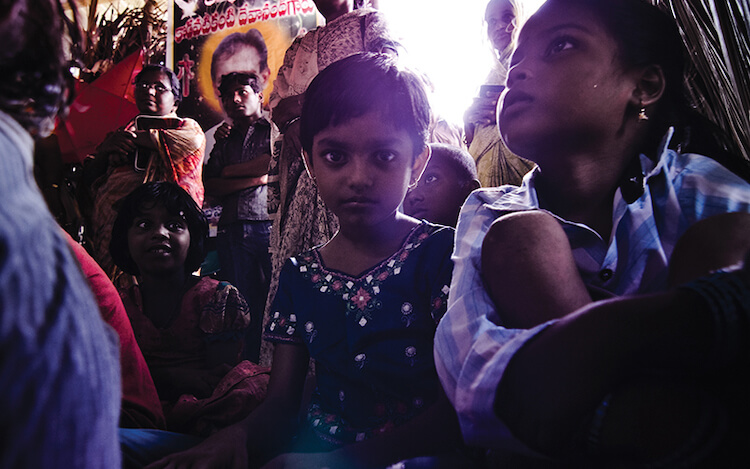 Crowd of people in a dimly lit space. The photo is a closeup on two young children. One is looking directly into the camera and one is looking up and to the side.
