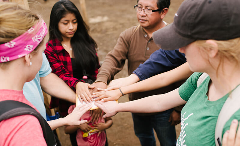 Group of young adults standing in a circle with their hands touching in the center.