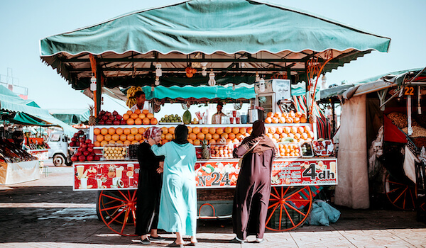 Large fruit stand with a teal cover over top. There are three patrons standing in front of the cart waiting to buy fruit.