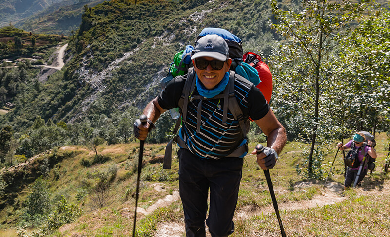 A man with hiking polls and a large backpack smiling as he climbs a mountain