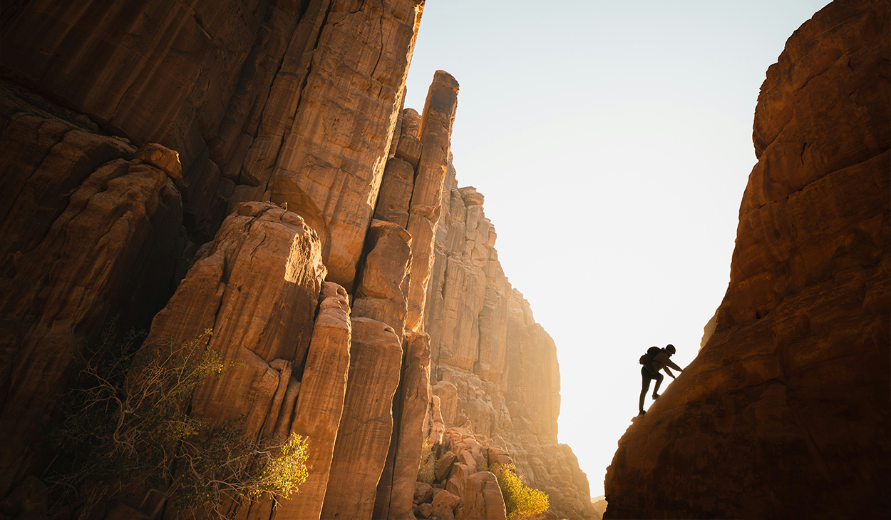 A person climbs up a rock face and is backlit by the sun in the background.