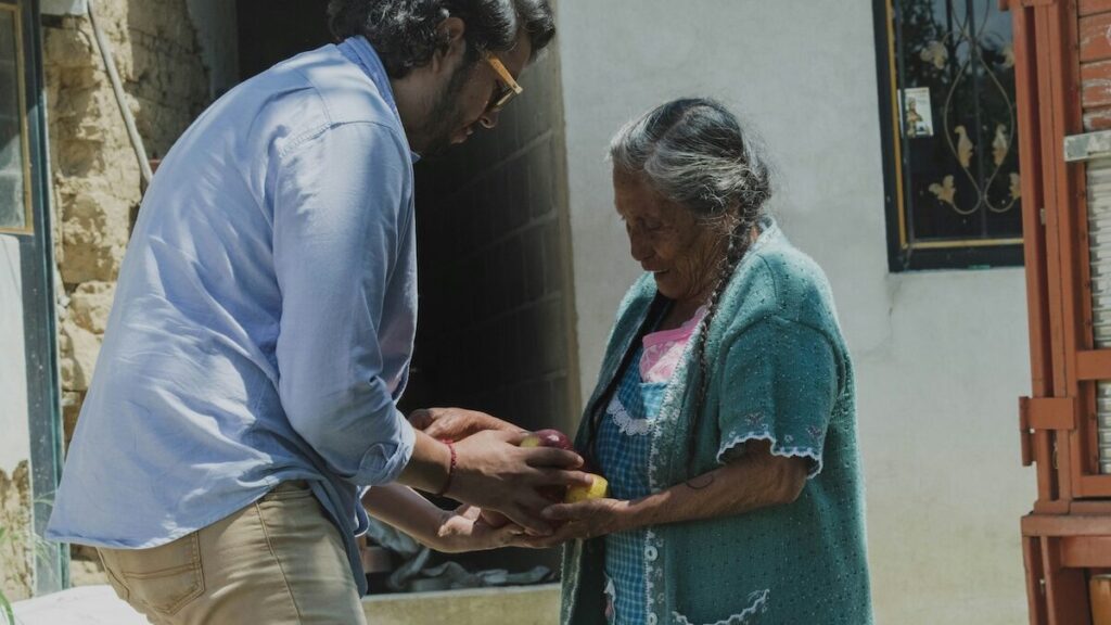 A man hands an object to an elderly woman outside, both focused on the exchange.