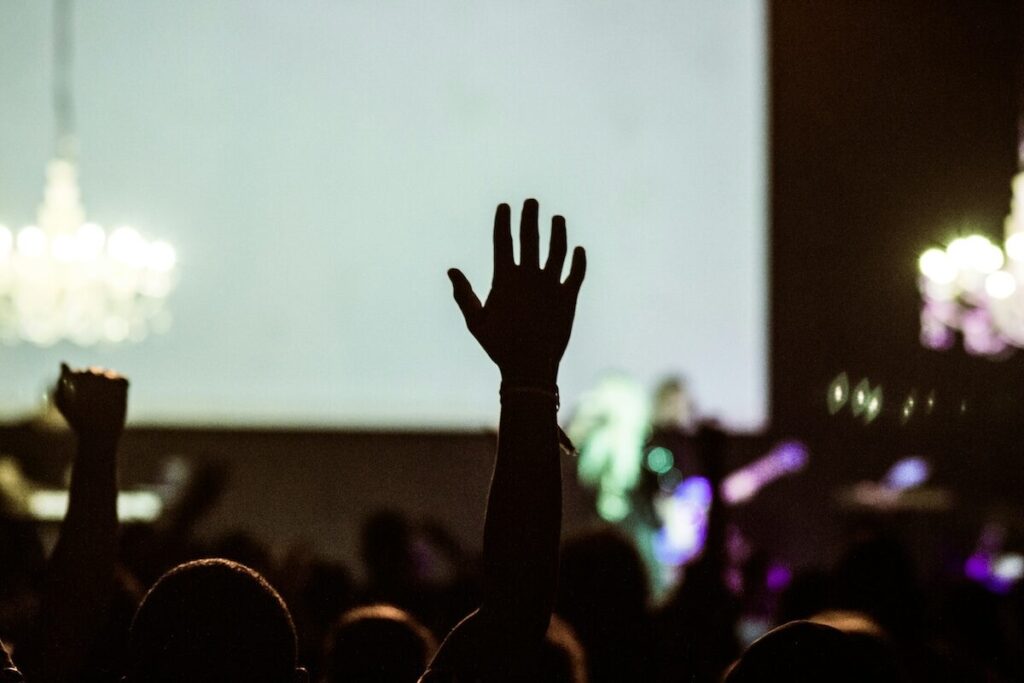 A raised hand in a crowd during a concert or live event, with stage lights and chandeliers visible in the background.